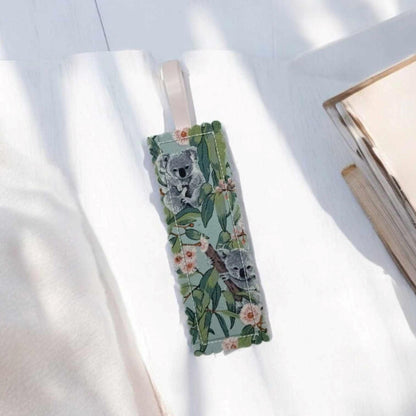 A green bookmark with koalas in a Eucalyptus tree with pink flowers. The bookmark has a pale pink ribbon loop attached.