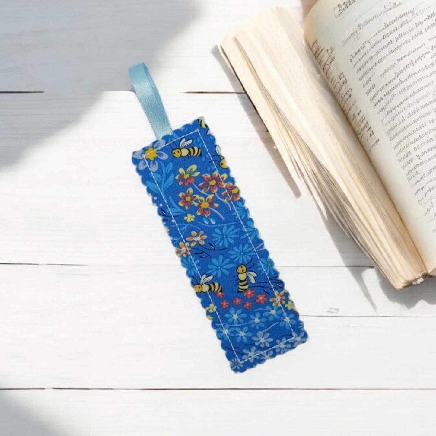 A blue bookmark with bees and flowers The bookmark has a pale blue ribbon loop attached.