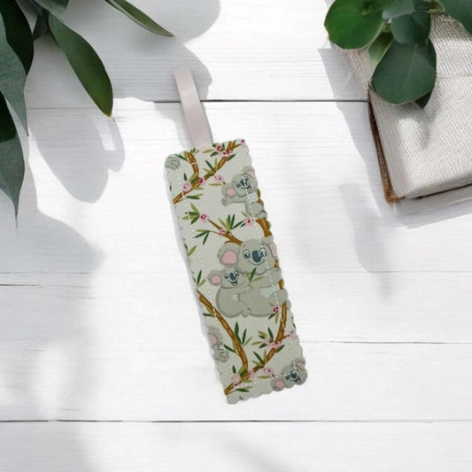 Handmade fabric bookmark featuring a cute koala on a white wooden surface. The koalas are climbing eucalyptus branches with pink flowers. The bookmark has scalloped edges & a pink ribbon loop. Props not included in sale.