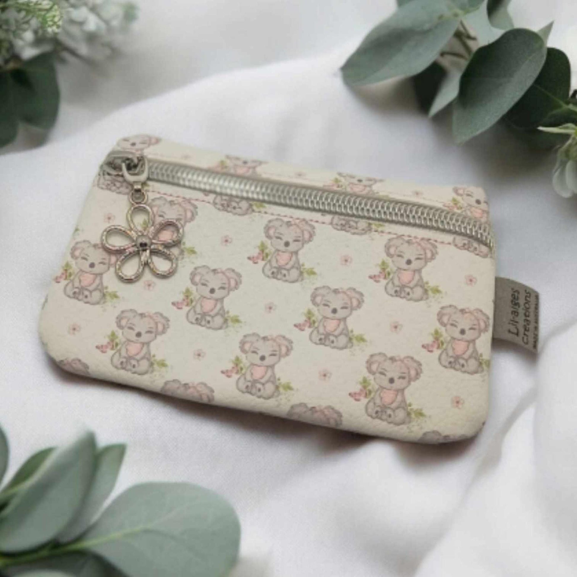 Koala themed vinyl coin purse with silver zip and flower zipper pull charm on a white piece of fabric with some green eucalyptus leaves and white flowers.