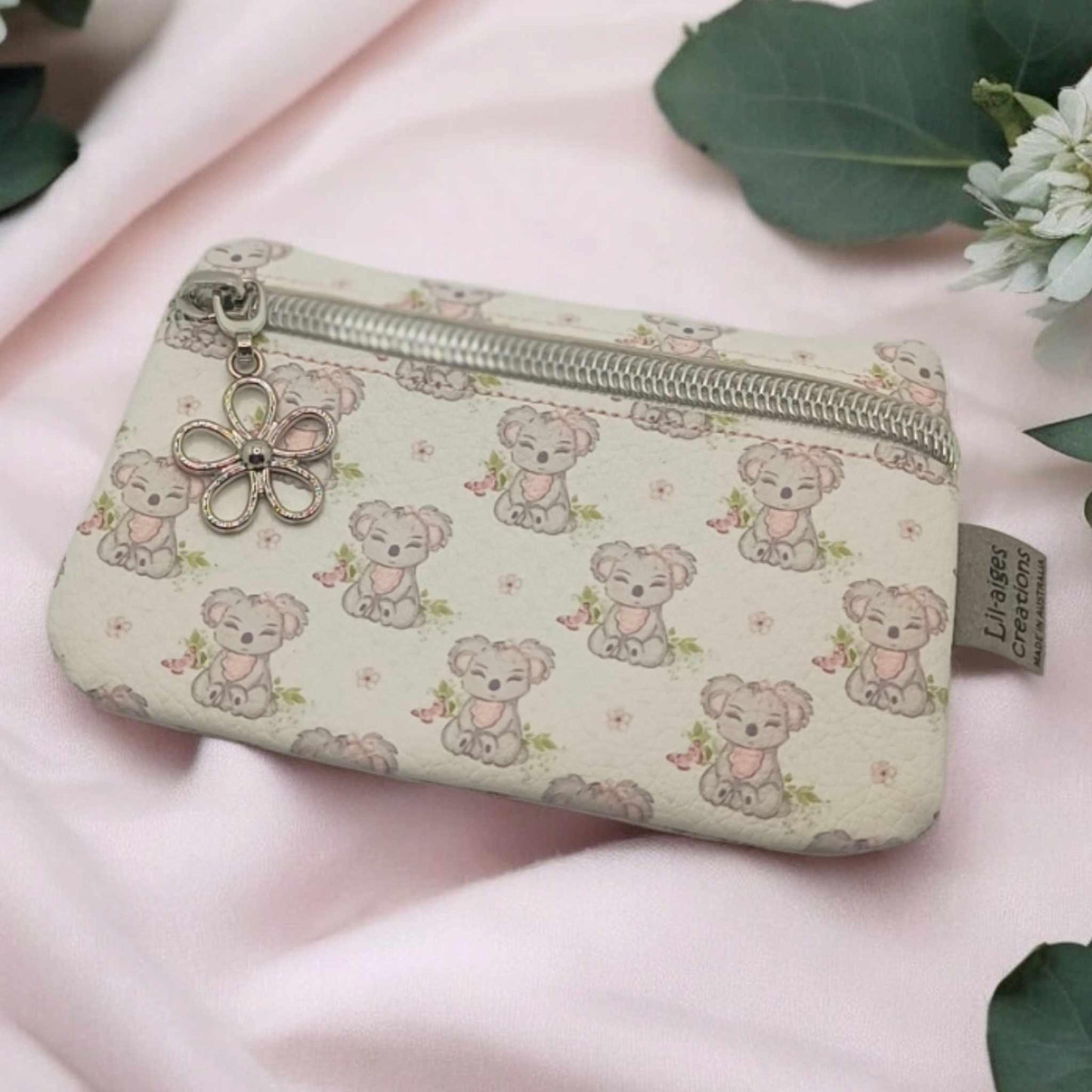 Koala themed vinyl coin purse with silver zip and flower zipper pull charm on a pale pink piece of fabric with some green eucalyptus leaves and white flowers.