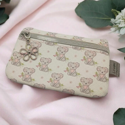Koala themed vinyl coin purse with silver zip and flower zipper pull charm on a pale pink piece of fabric with some green eucalyptus leaves and white flowers.