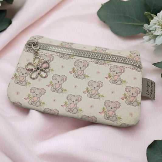 Koala themed vinyl coin purse with silver zip and flower zipper pull charm on a pale pink piece of fabric with some green eucalyptus leaves and white flowers.
